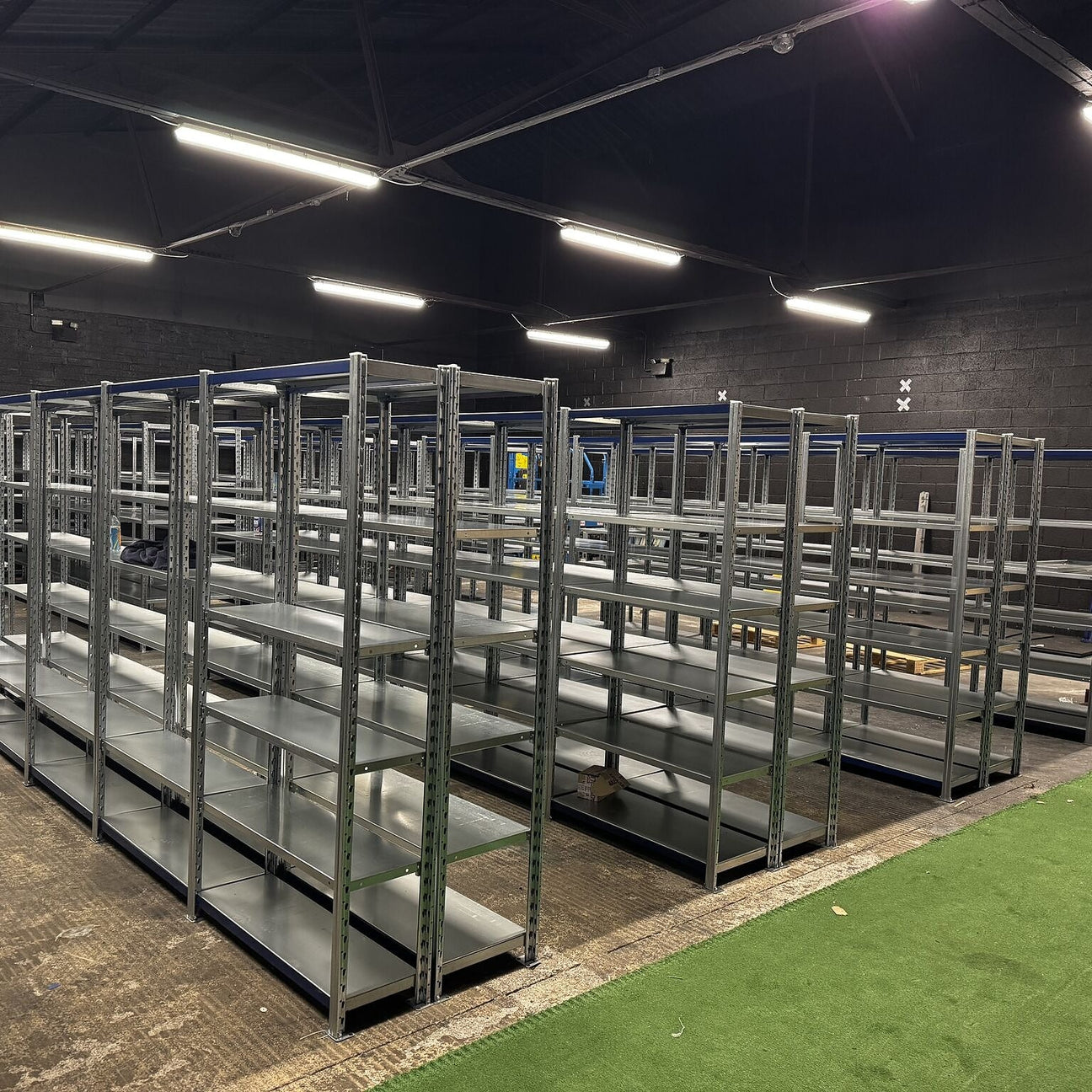 Rows of empty metal shelving units are arranged in a large, industrial-style room with concrete floors and overhead lighting.
