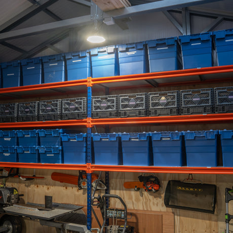 Shelving unit in a workshop, holding large blue storage bins and black plastic crates. Workshop tools and equipment are visible below the shelves.