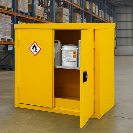 A yellow safety cabinet with a flammable materials warning label stands open in a warehouse, showing chemical containers on the shelf inside.