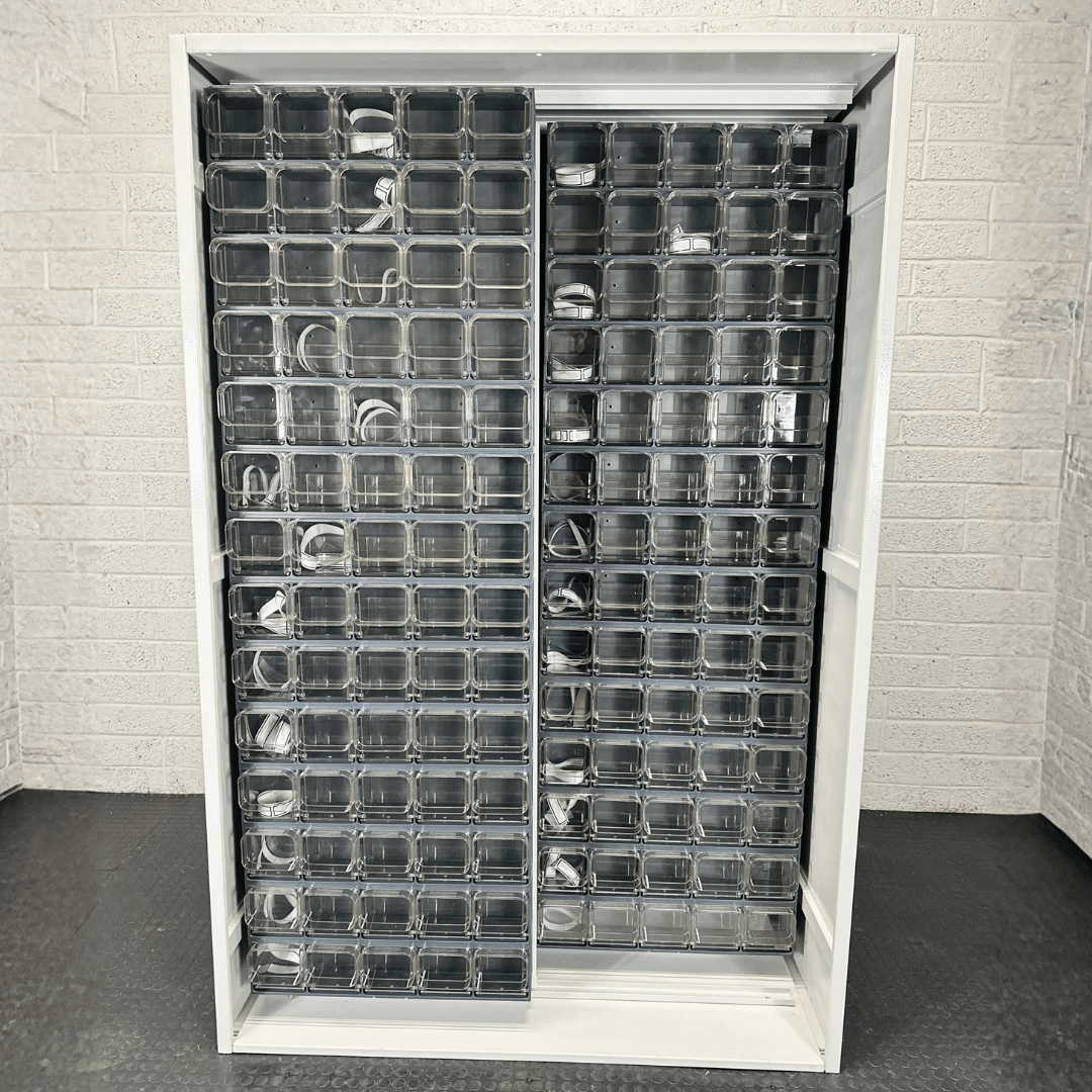 A white shelving unit with multiple clear plastic compartments, some containing coiled cables, stands against a light-colored brick wall.