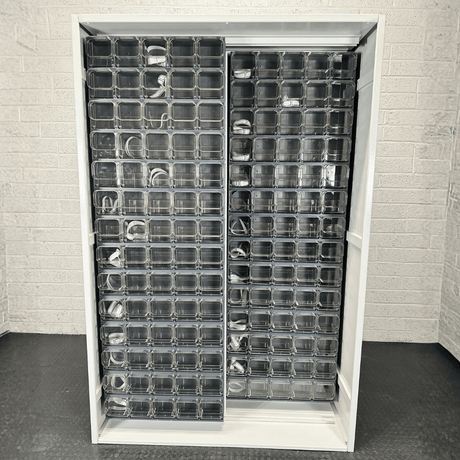 A white shelving unit with multiple clear plastic compartments, some containing coiled cables, stands against a light-colored brick wall.