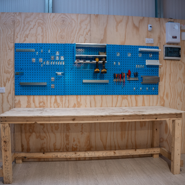 A wooden workbench stands in front of a wall-mounted blue pegboard holding assorted tools and storage bins in a workshop.