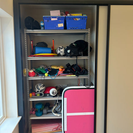 A metal shelf in a closet holds sports balls, toys, fitness equipment, bags, a blue crate, and a pink foldable mat, with various items stacked and stored on each shelf.