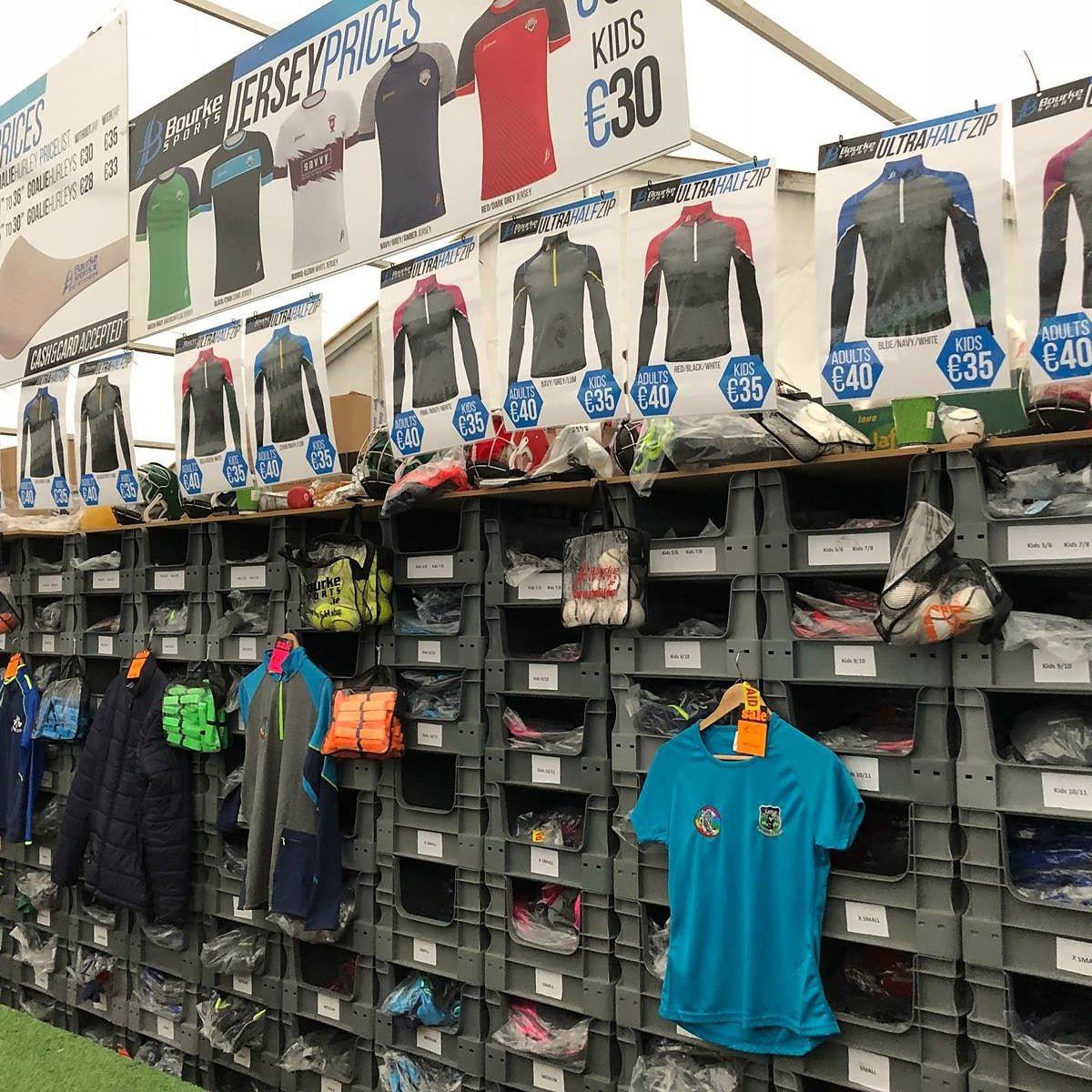 Shelves and bins display various sports jerseys, shirts, and gear for sale under signs listing adult and kids’ prices at a retail booth.