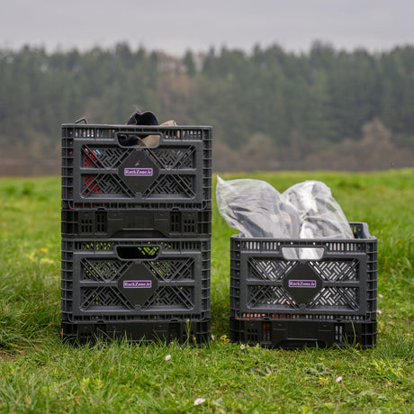 Four black plastic crates containing various items are stacked on grass outdoors, with a forest visible in the background.