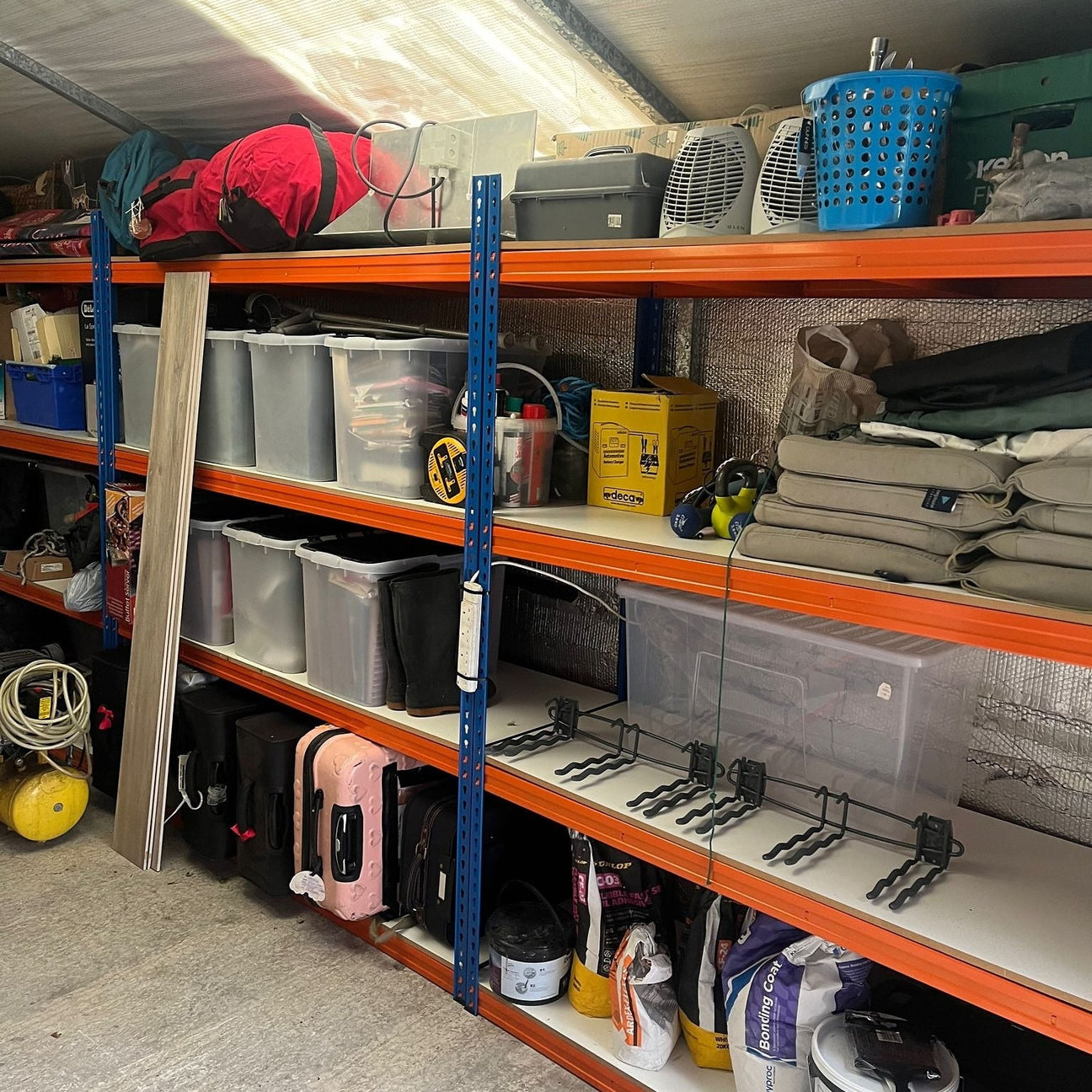 Metal shelving unit in a storage area holds various items including plastic bins, luggage, camping gear, a folded table, and baskets.