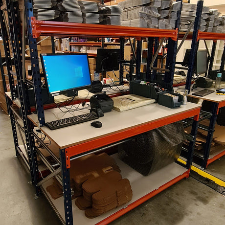 A warehouse packing station with a computer, keyboard, printer, and packing materials including boxes, bubble wrap, and tape, organized on shelves.