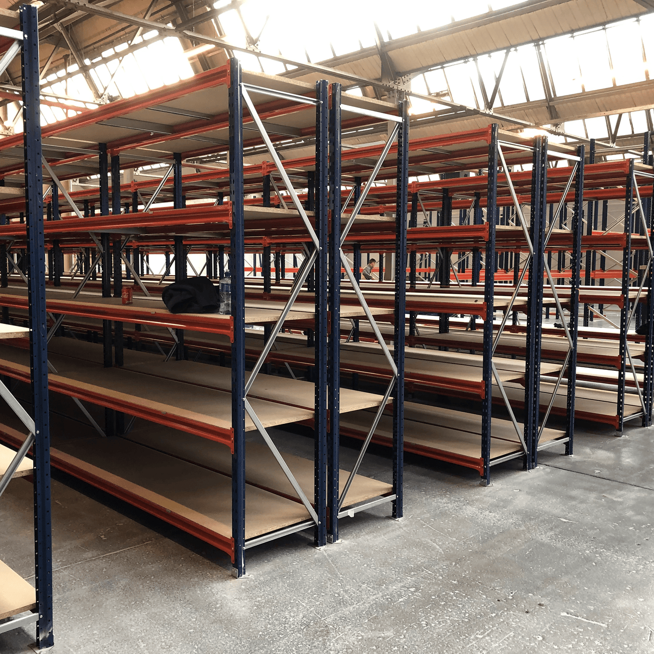Rows of empty industrial metal shelving units are arranged inside a large warehouse with high windows and a concrete floor.