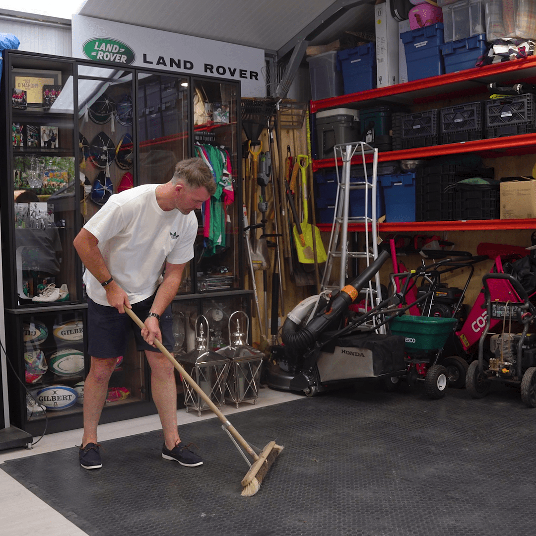 A man in a white shirt and shorts sweeps the floor of a garage filled with tools, storage bins, and gardening equipment.
