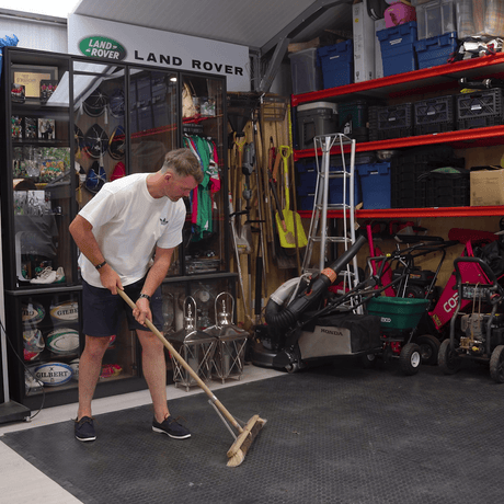 A man in a white shirt and shorts sweeps the floor of a garage filled with tools, storage bins, and gardening equipment.