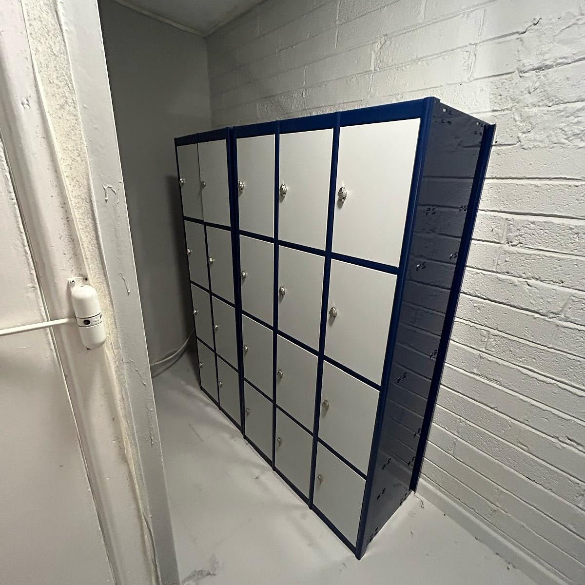 A set of blue and white lockers is placed against a white brick wall in a small, empty room with a concrete floor.