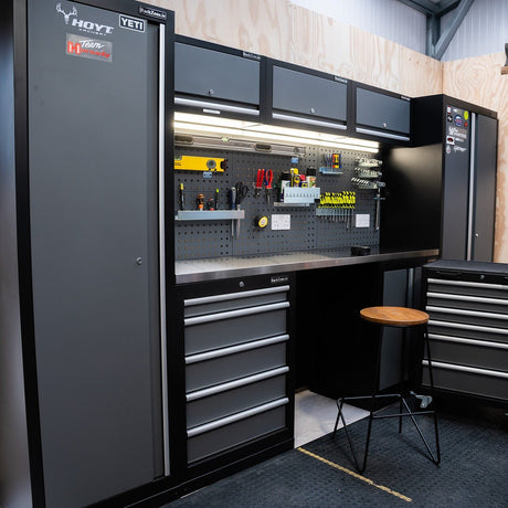 A workshop with black and gray cabinets, drawers, a pegboard with organized tools, a workbench, and a wooden stool.