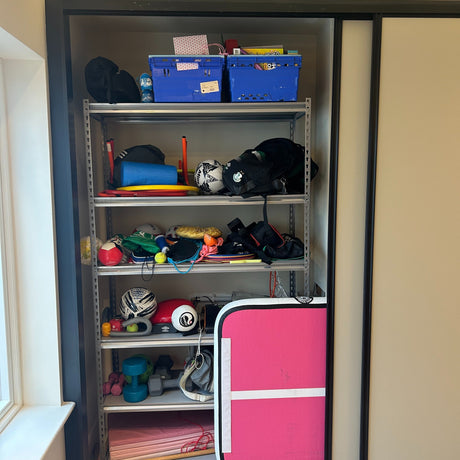 A metal shelf in a closet holds sports balls, toys, fitness equipment, bags, a blue crate, and a pink foldable mat, with various items stacked and stored on each shelf.