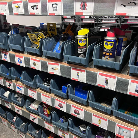 Shelves display various hardware items, including WD-40 cans, safety glasses, tape, and tools, organized in plastic bins with labeled product cards below each item.