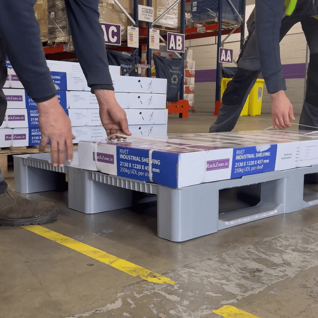 Two people load boxes labeled "industrial shelving" onto a gray plastic pallet in a warehouse with labeled aisles and stacks of boxes in the background.