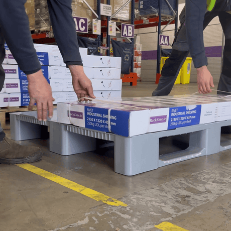 Two people load boxes labeled "industrial shelving" onto a gray plastic pallet in a warehouse with labeled aisles and stacks of boxes in the background.