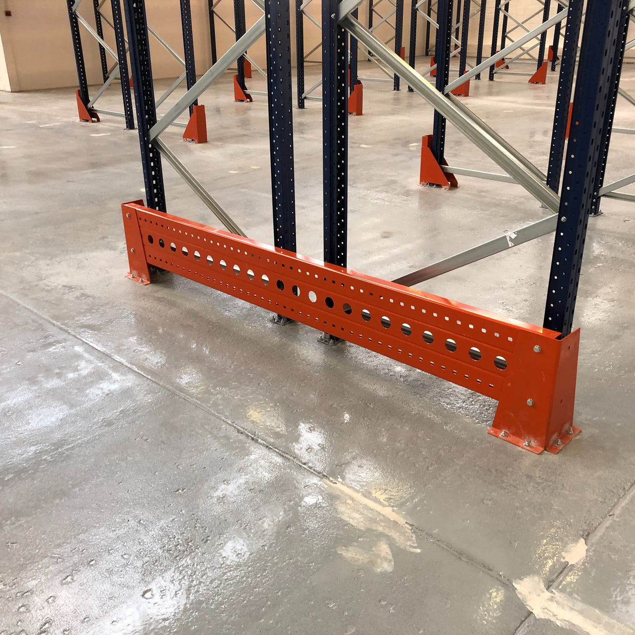 A metal rack guardrail with multiple holes is bolted to a warehouse floor, protecting the lower part of a pallet racking system.