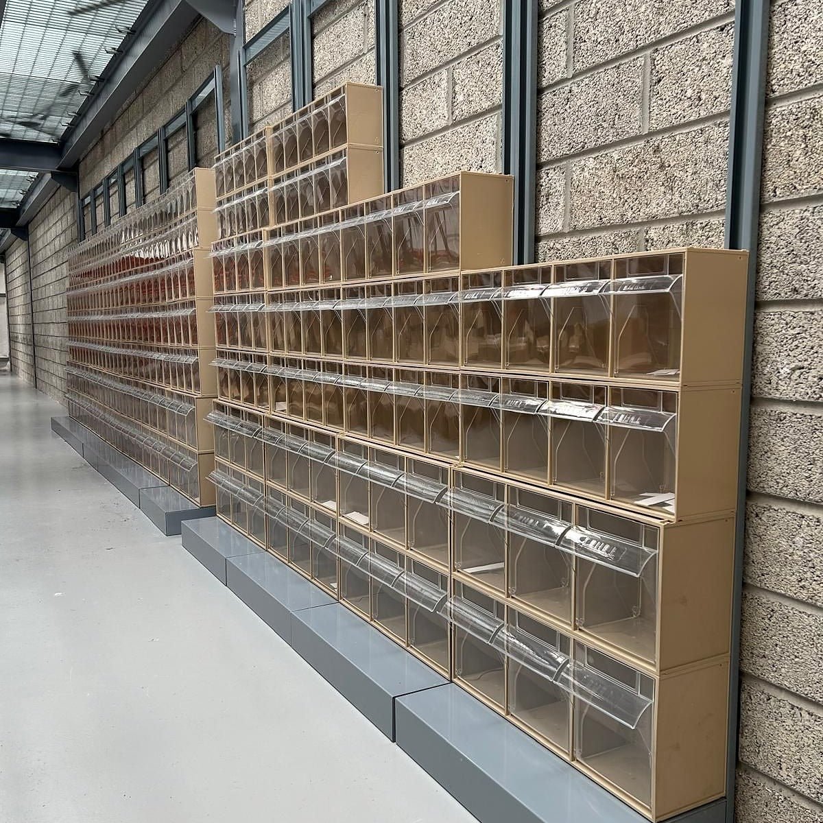 A long row of empty, clear-fronted storage bins arranged against a brick wall in a hallway with a concrete floor.