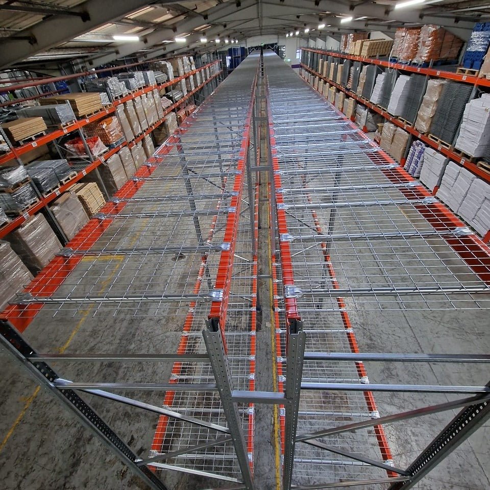 View of empty metal shelving in the center aisle of a warehouse, with stacked boxes and goods on shelves along both sides.