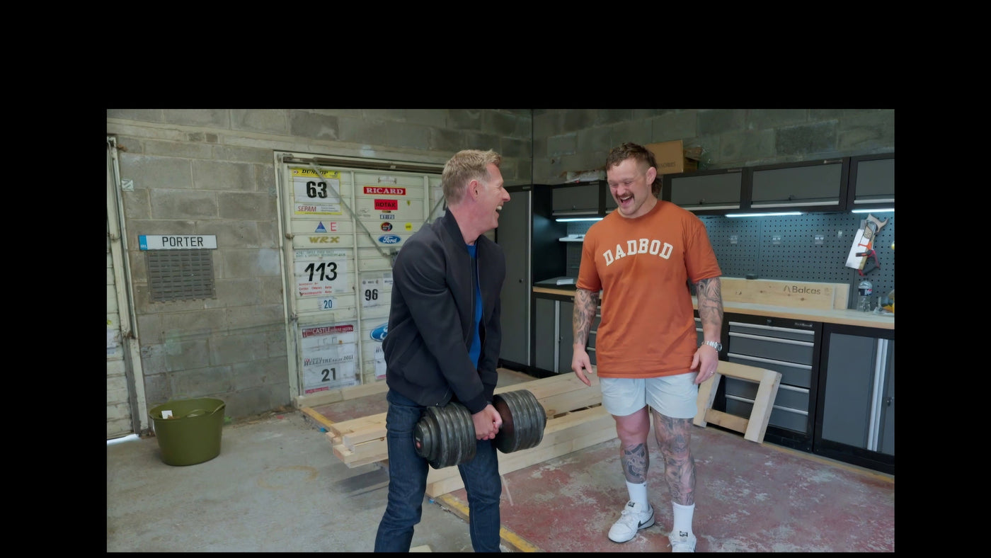 Two men in a workshop setting, one lifting weights and the other observing.
